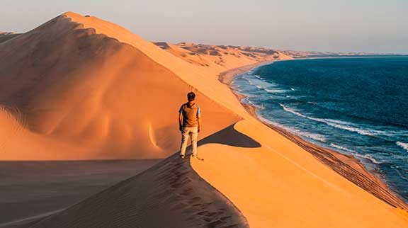 Mann steht auf roten Dünen einer Wüste, daneben sieht er das Meer.
