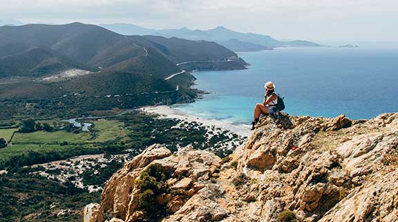 Frau sitzt auf Berg und schaut auf Korsika mit Strand und Bergen hinunter.