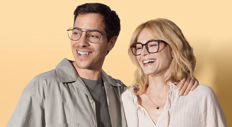 Smiling man in dark, double-bridged glasses with his arm around a smiling woman in dark, rectangular glasses, against a yellow background