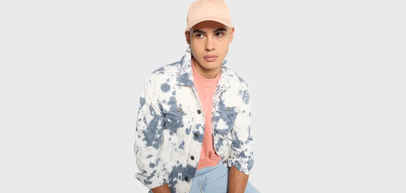 Junger Mann mit einer rosa Kappe, einem rosa Hemd und einer blau-weißen Batikjacke, der mit den Händen auf seinen Knien sitzt, vor einem schlichten Hintergrund.