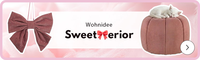 Rechts liegt eine schlafende weiße Katze auf einem rosafarbenen, runden Pouf. Links hängt eine große, rosafarbene Stoffschleife mit kleinen weißen Punkten. In der Mitte steht der Text 'Wohnidee Sweet erior', mit einer pinken Schleife in der Mitte. Der Hintergrund ist in sanften Rosatönen gehalten. Unten rechts befindet sich ein einzelner, weißer Pfeil in einem Kreis.