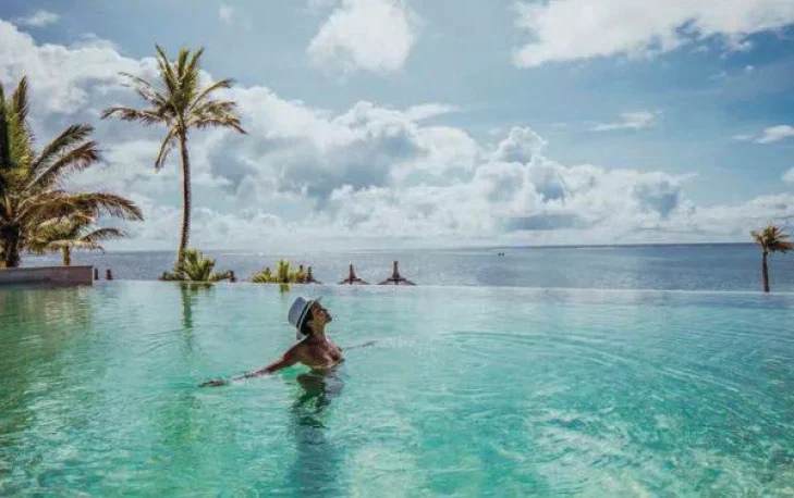A lady basks in the glow of the sun and she enjoys bathing in an infinity pool with the ocean in the horizon.