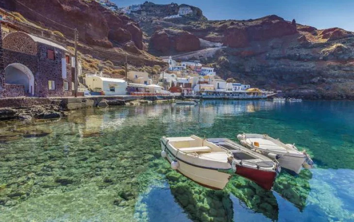 Small boats floating on clear turquoise water in a peaceful harbour in Santorini, with whitewashed houses and rugged volcanic cliffs in the background.