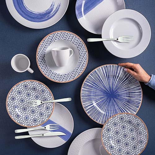 A collection of ceramic plates with various blue and white geometric and abstract patterns, arranged on a dark blue background. A hand is holding the largest plate featuring a radial line design. A white cup and several forks are placed across the arrangement, emphasising the aesthetic display.