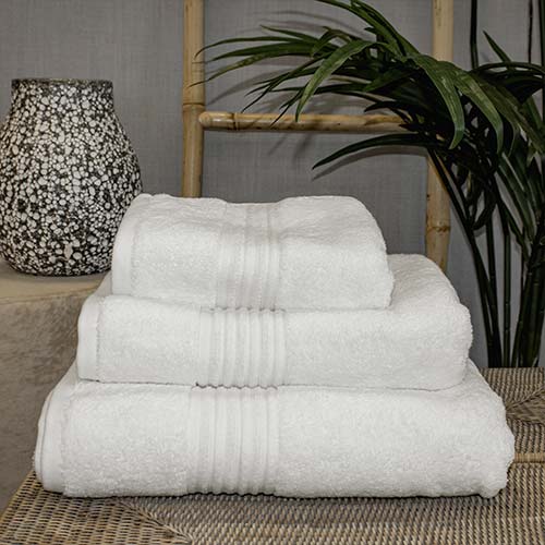 A stack of three folded white towels with subtle striped patterns, placed on a woven surface. In the background, there is a decorative black and white speckled vase and a lush green plant.