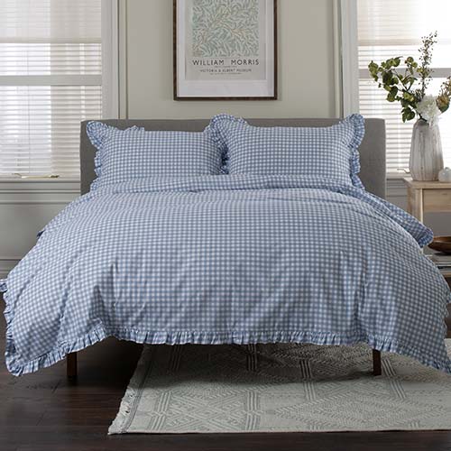 A neatly made bed features blue and white gingham bedding with ruffled edges. Two matching pillows rest against a grey upholstered headboard. A framed William Morris print hangs on the wall above, and a vase with flowers sits on a bedside table to the right. Blinds cover the windows, filtering soft 