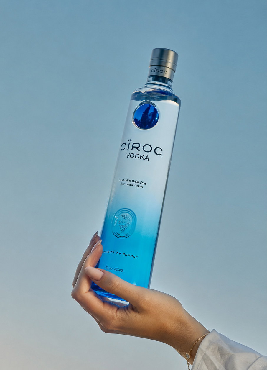 Hand holding a Cîroc Vodka bottle with blue gradient glass against a pale blue background, product label visible