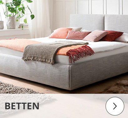 Kategorie Schlafzimmer: Helles Schlafzimmer mit einem großen Boxspringbett in hellgrauem Stoffbezug. Das Bett ist dekoriert mit Decken und Kissen in Braun- und Rottönen. Im Hintergrund steht eine Grünpflanze vor einem Fenster. Der Schriftzug 'BETTEN' ist unten eingeblendet.