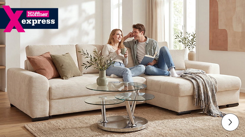 Ein großes, helles Ecksofa mit strukturierter Oberfläche steht in einem modern eingerichteten Wohnzimmer. Zwei Personen sitzen entspannt nebeneinander auf dem Sofa, eine Person hält ein Buch, die andere eine Tasse. Vor dem Sofa steht ein runder, dreistufiger Glastisch mit Metallgestell, darauf eine Vase mit Zweigen. Im Hintergrund sind große Fenster mit hellen Vorhängen und ein abstraktes Wandbild zu sehen. Links oben befindet sich ein auffälliges, mehrfarbiges Logo mit dem Schriftzug 'Höffner express'.