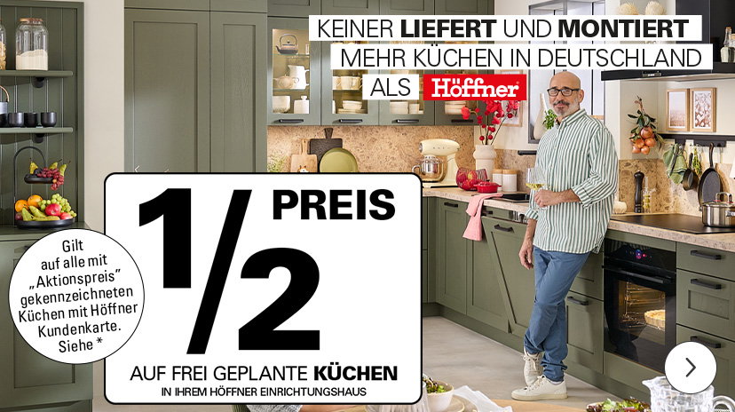 Eine moderne Einbauküche mit grünlichen Fronten, schwarzen Griffen und integrierten Elektrogeräten ist zu sehen. Auf der Arbeitsplatte stehen verschiedene Küchenutensilien. Im Hintergrund sind offene Regale mit Geschirr und Dekorationen. Eine Person steht an der Küchenzeile. Im Vordergrund befindet sich ein großes, weißes Werbeschild mit folgender Info: ‚½ PREIS AUF FREI GEPLANTE KÜCHEN IN IHREM HÖFFNER EINRICHTUNGSHAUS‘. Weiter sagt die Grafik, dass die Aktion auf ‚Aktionspreis‘-Artikel gilt und nur mit Kundenkarte.