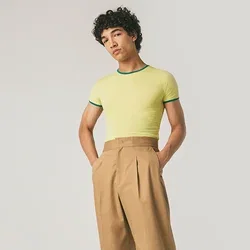 Joven de pie con las manos en los bolsillos, vistiendo una camiseta ajustada amarilla, pantalones marrones anchos y zapatos oscuros, frente a un fondo simple.