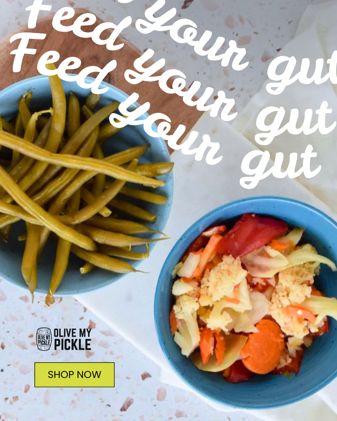Two blue bowls of green beans and mixed pickled vegetables on a speckled counter. White text "Feed your gut" is overlaid, with an "Olive My Pickle" logo and "SH