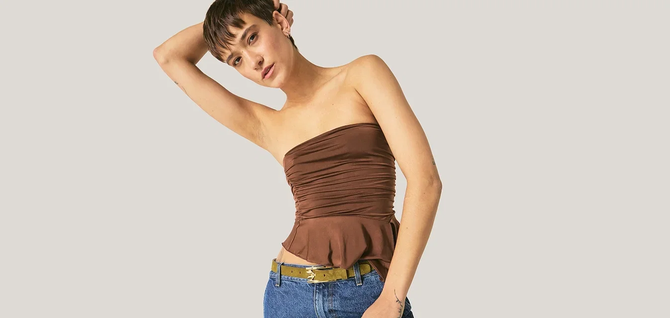 Vrouw met een bruin strapless topje en blauwe jeans leunt naar één kant, waarbij ze één arm op haar hoofd laat rusten tegen een effen achtergrond.