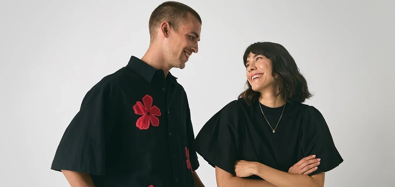 Man en vrouw glimlachend en naar elkaar gewend, man in zwart shirt met rode bloemen, vrouw in zwart top en witte rok tegen een effen achtergrond.