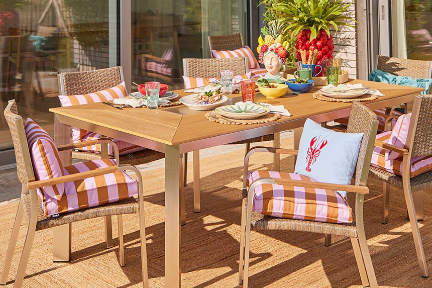 Das Bild zeigt eine moderne Terrasse mit einem rechteckigen Holztisch und sechs Stühlen, die mit gestreiften Sitzkissen bezogen sind. Auf dem Tisch stehen bunte Dekorationen, Geschirr und Gläser.