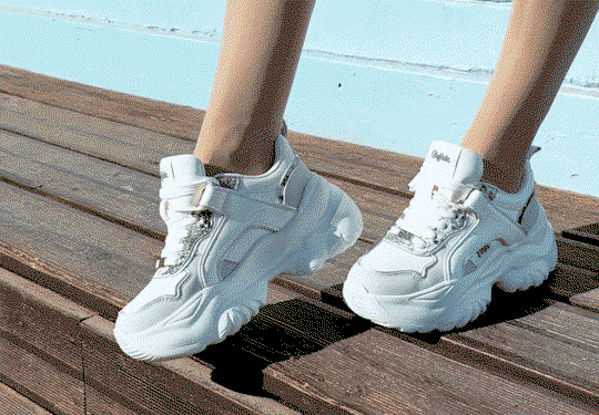 White chunky sneakers with decorative details worn on a wooden boardwalk against a light blue wall