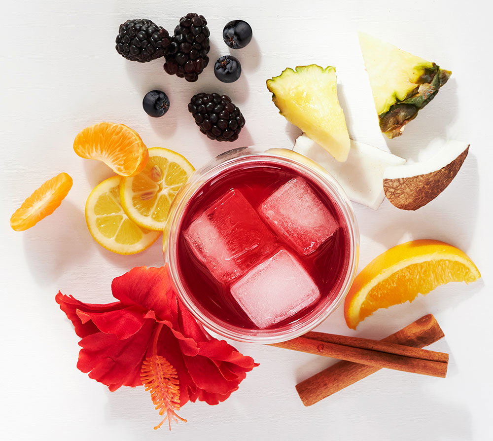 Hibiscus tea in a teacup surrounded by fruits: yuzu, pineapple pieces, coconut pieces, blackberries, blueberries, orange slice, cinnamon sticks, tangerines and a hibiscus flower.