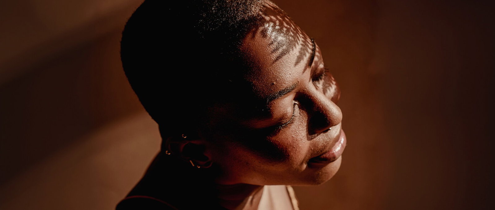 Woman with eyes closed, face tilted upward in natural light with leaf shadow patterns across her skin