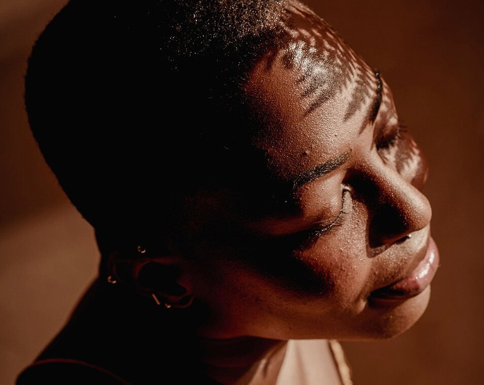 Woman with eyes closed, face tilted upward in natural light with leaf shadow patterns across her skin
