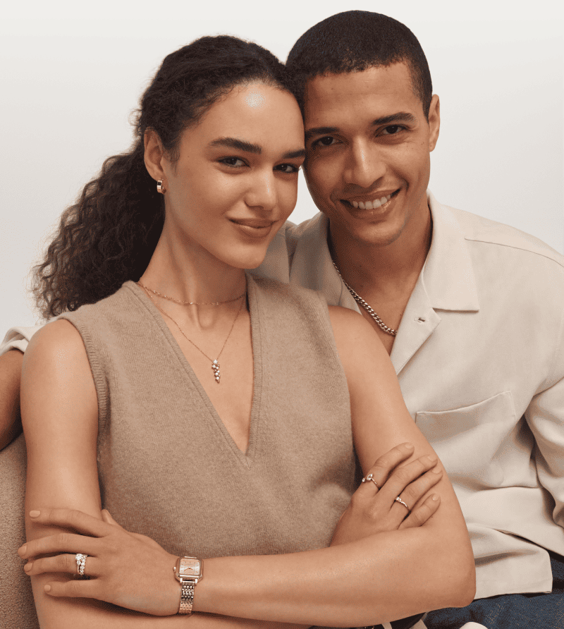 Smiling couple wearing jewelry. Woman is wearing layered necklaces, rings and a watch. Man is wearing a chain necklace.