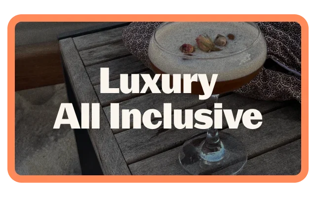 Coral-orange border, 'Luxury All Inclusive white bold text, overhead view, dark wooden tray with grooves, white plate with nuts or dried fruits, wine glass with blue liquid, ornate patterned textile and rounded corners.