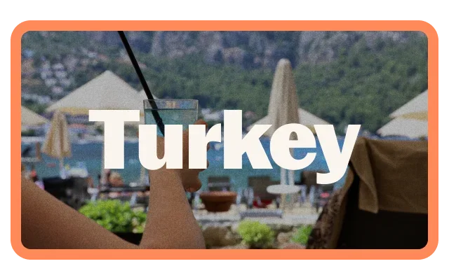 Coral-orange border, 'Turkey' white bold text, blurred Turkish architecture background, conical rooftops, hillside with green vegetation, outdoor café with umbrellas, earth tones and rounded corners.