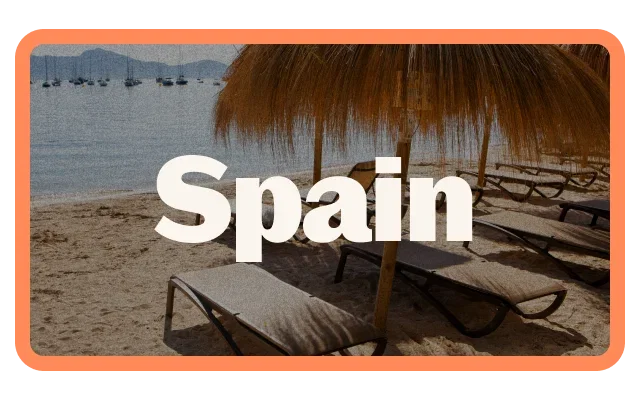 Coral-orange border, 'Spain' white bold text, beach scene, blue-grey water, anchored boats, misty mountains, beige sand, two beach loungers, thatched palapa umbrella, golden-brown palm fronds, wooden pole and dappled shadows.