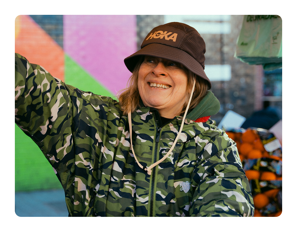 Person wearing a HOKA bucket hat smiling, in a market stall with oranges.