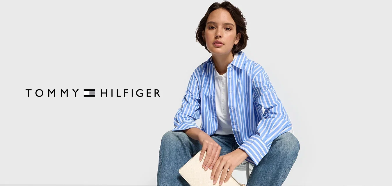 Blauw-wit gestreepte blouse met lange mouwen, witte t-shirt, lichtblauwe jeans en witte sneakers. Houdt een crèmekleurige clutch met een logo vast.