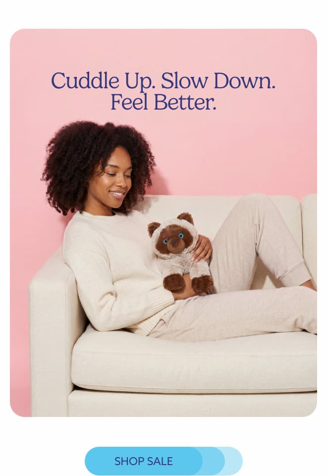 A woman relaxing on a couch with Siamese Cat Warmies. Shop Sale.