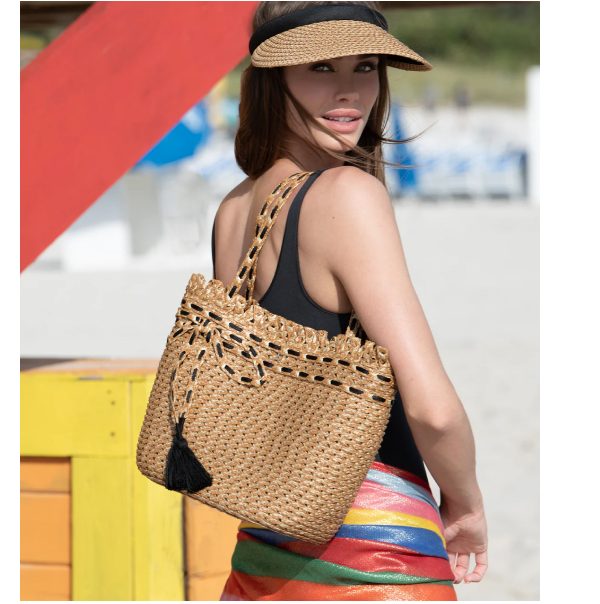 A woman wearing a black swimsuit and visor, holding a straw tote bag on the beach.