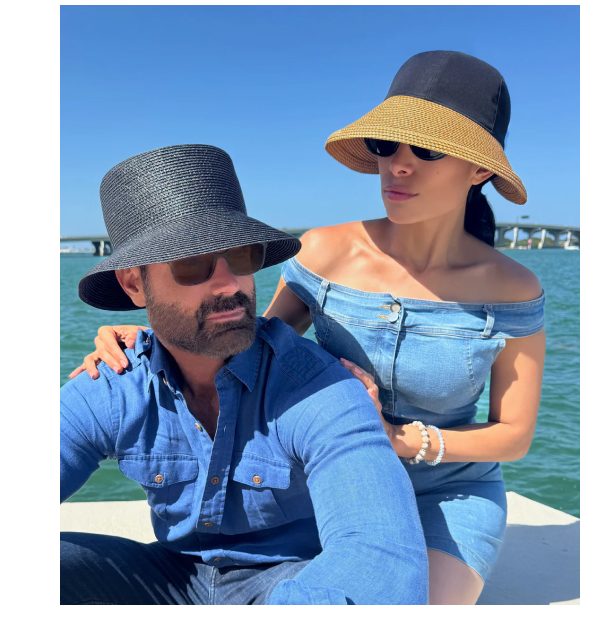 A couple wearing hats and sunglasses by the water, with a bridge in the background.