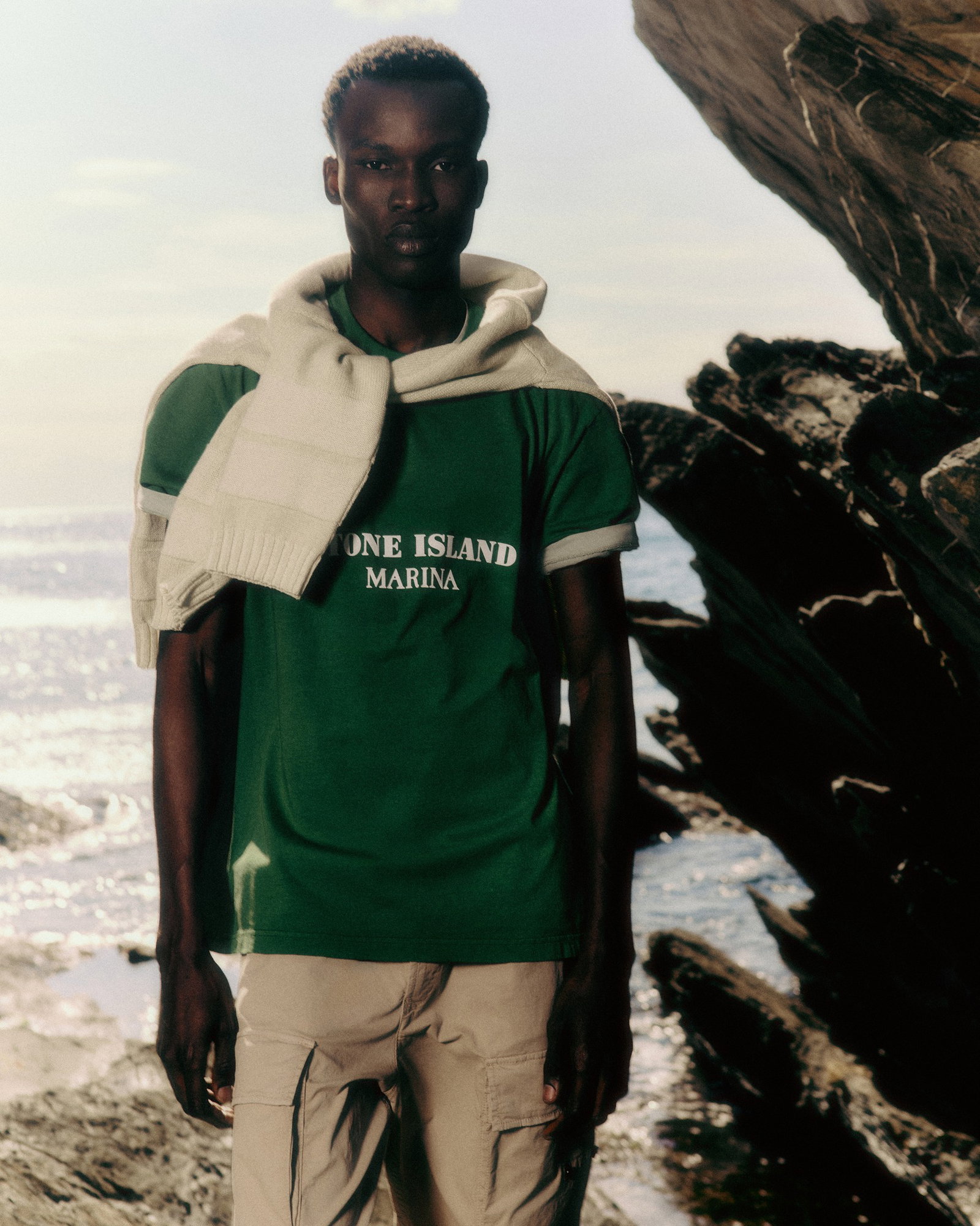 Man wearing green 'One Island Marina' t-shirt with sweater over shoulders standing by a rocky seaside
