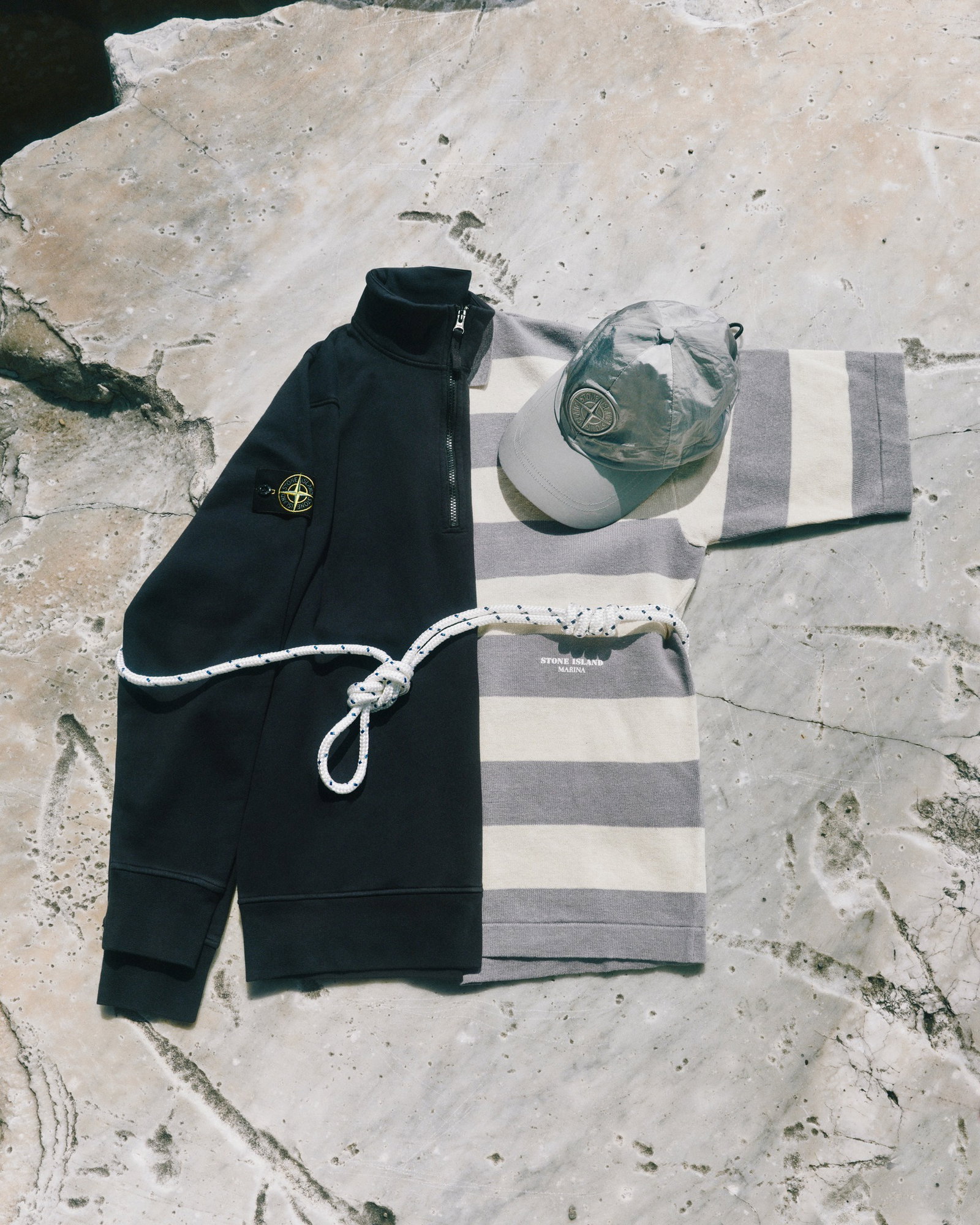 Flat-lay of navy half-zip jumper with compass badge, grey-and-cream striped long-sleeve top, grey cap with logo and knotted white rope on pale rock