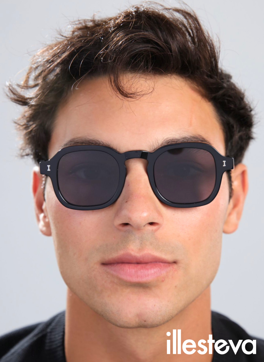 A close-up portrait of a man wearing black, slightly angular rounded sunglasses with the white illesteva logo printed on the right.
