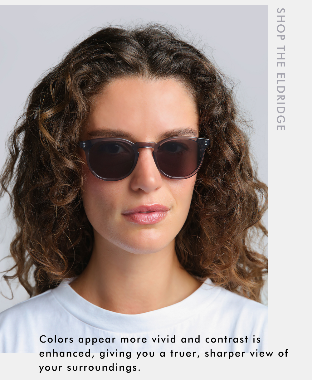 A promotional headshot of a woman with curly brown hair looking straight ahead while wearing the Eldridge sunglasses in a translucent grey frame with dark lenses. Vertical text along the top right reads, 