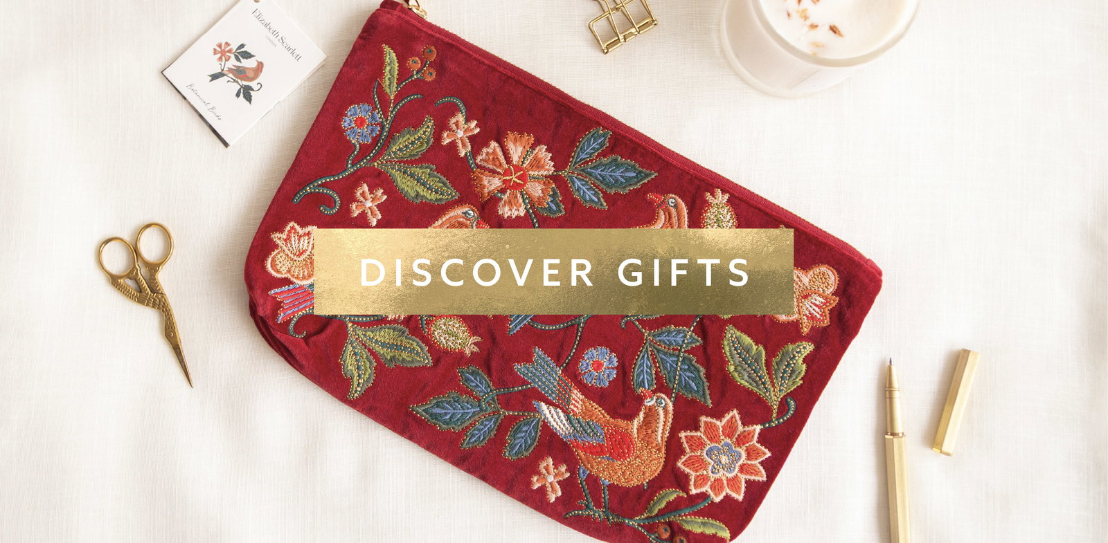 Luxury embroidered floral pouch by Elizabeth Scarlett styled on a cream linen desk with gold scissors, candle and stationery accessories alongside a gold discover gifts banner.