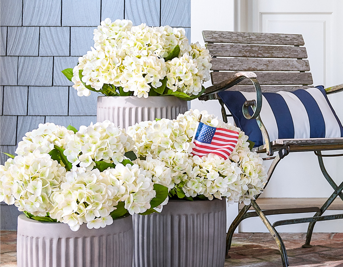 White hydrangeas in gray planters beside a rustic chair with a patriotic pillow and mini American flag.