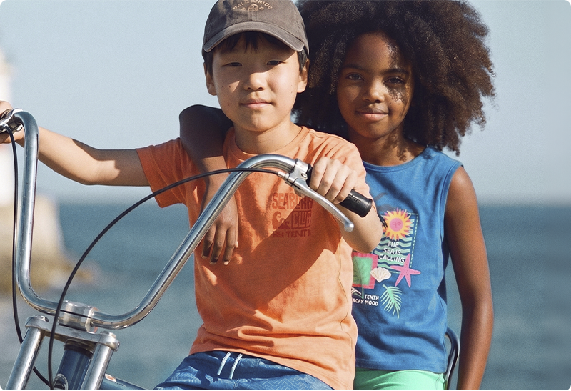 Two smiling children—boy wearing cap steering a bicycle and girl with curly hair hugging him—by the seaside on a sunny day