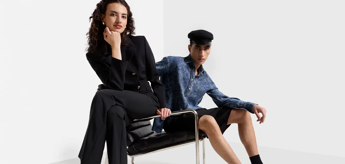 Deux mannequins posent dans un cadre minimaliste. L'un porte un costume noir et des escarpins noirs à lanières ; l'autre porte une chemise bleue et un short noir, assis.