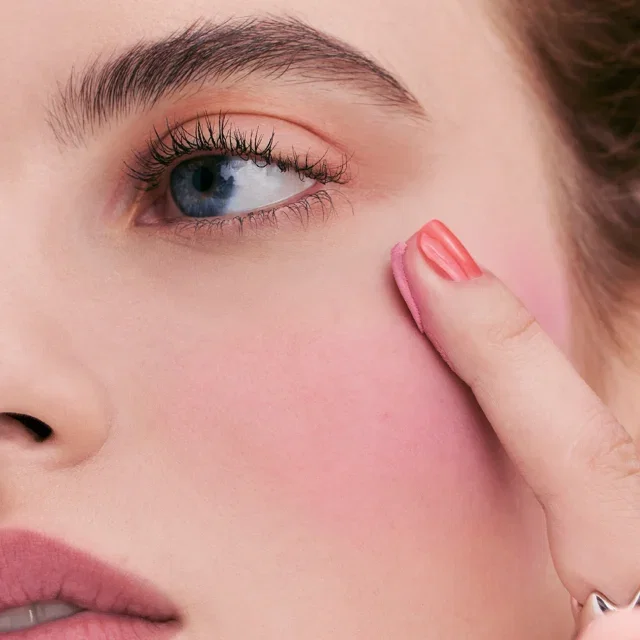 Close-up of a woman's face and finger applying Pot Rouge onto her cheek.