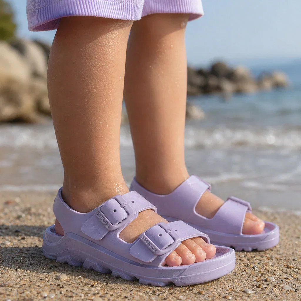 Child wearing lavender EVA sandals with buckle straps on a sandy beach with ocean in background.