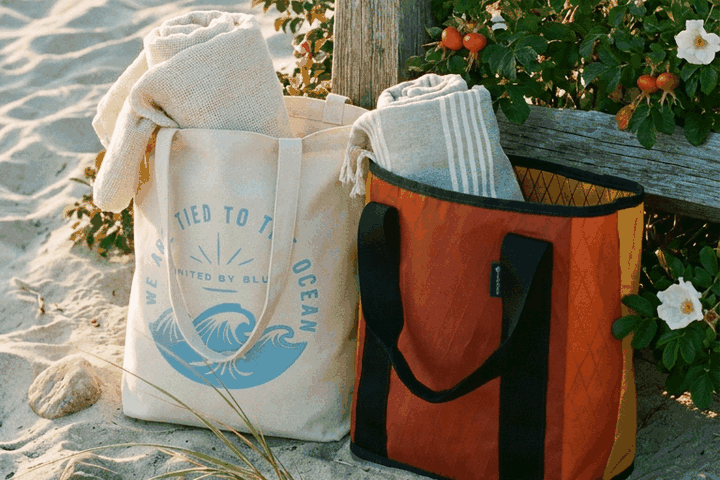 The Westward Tote and My Favorite Daily Tote resting against a weathered seaside fence with beach roses in bloom