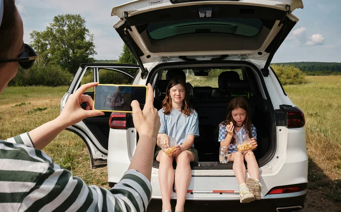 A mother is shown taking a photo of her girls sitting in the back of a car