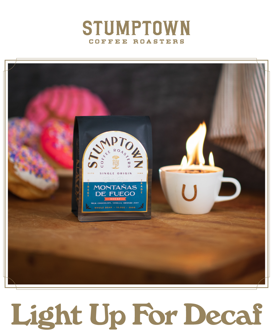 Stumptown single origin Montaña de Fuego decaf coffee bag displayed on a wooden table beside a white coffee cup with a horseshoe logo and flames rising from the cup. Colorful sprinkled donuts sit blurred in the background. Headline reads: “Light Up For Decaf.”