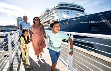 People walk along a cruise ship deck, large ship behind, bright sunny day, metal railings, casual summer clothing.