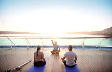 Three people sit on mats facing an instructor on a deck, practicing yoga by the ocean at sunrise.