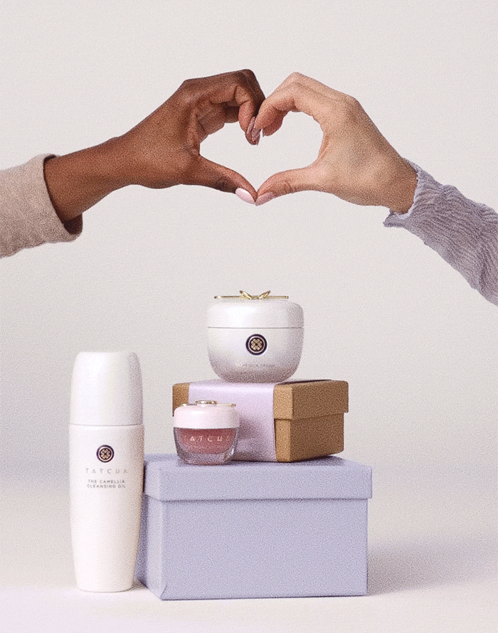 An assortment of Tatcha products, including the Camellia Cleansing Oil, Silk Cream, and Kissu Lip Mask stacked on some gift boxes, while two hands make a heart symbol.
