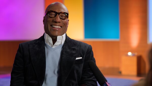  Byron Allen smiles as he sits by a mic for an interview. He is wearing a dark suit coat, a light blue vest and a white collared shirt. 
