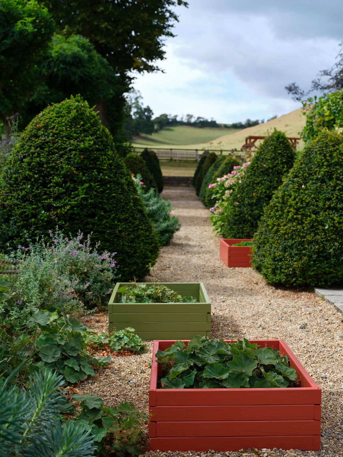 Gravel garden path lined with clipped greenery and raised planters painted red and green against a countryside backdrop.